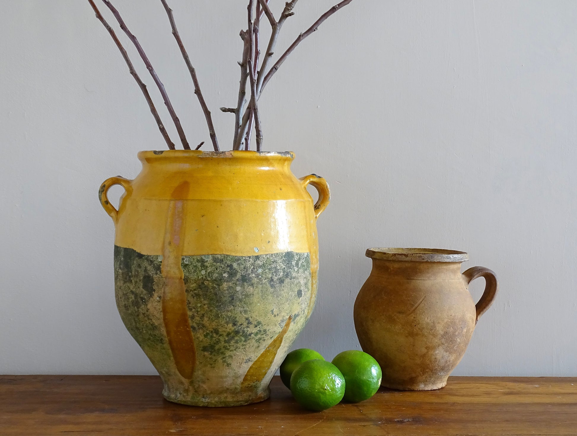 French antique confit pot with yellow glaze filled with branches, alongside a terracotta jug and fresh limes
