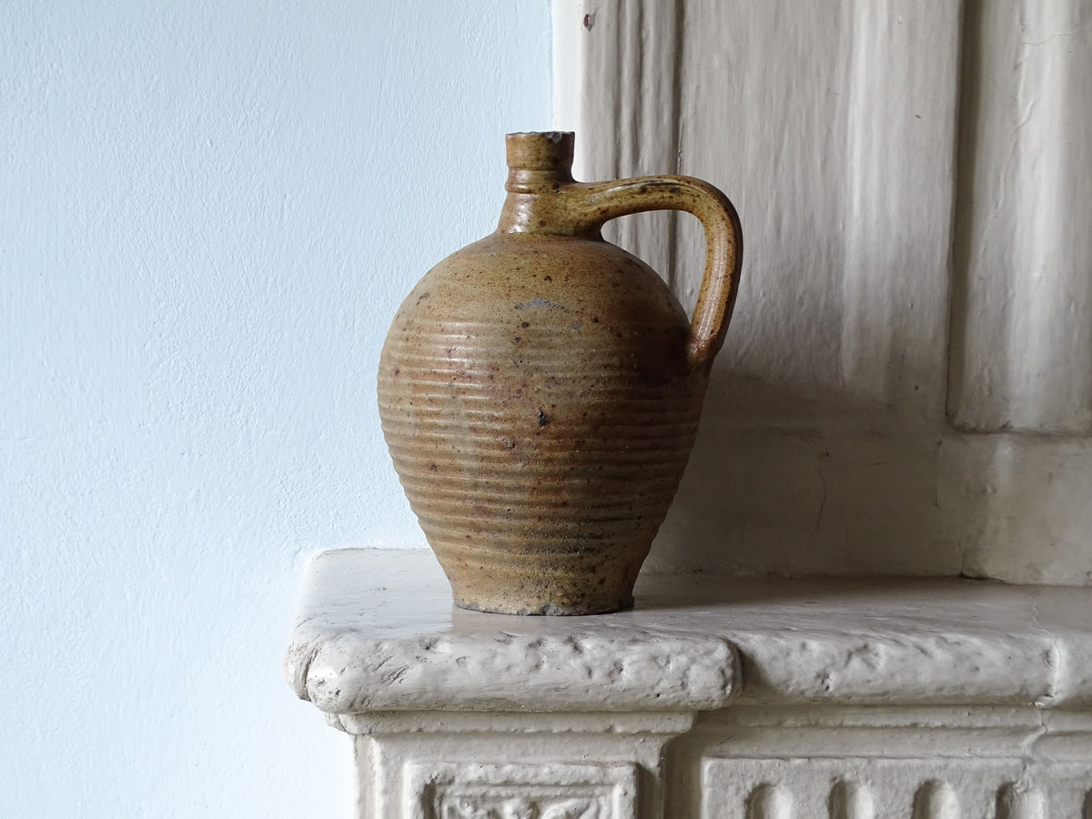 A rustic French antique ceramic jug with a ribbed body and handle, featuring a brown and beige speckled glaze, displayed on a white stone mantelpiece against a light blue wall.