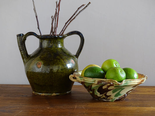 French antique orjol pitcher with a dark green glaze filled with branches. Alongside a French antique ceramic bowl with green, yellow and brown glaze filled with fresh limes.