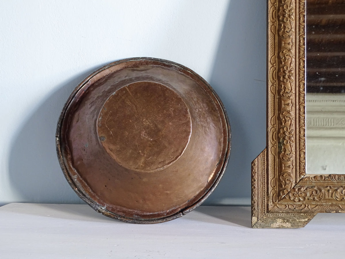 Underside of an antique copper basin with rustic finish next to a gold mirror
