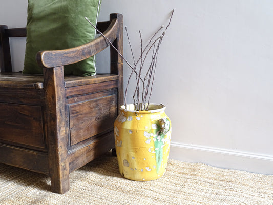 Large French antique ceramic jar or vinaigrier with a confit pot yellow glaze with green detailing. It is filled with branches and sits on a natural jute rug, next to an antique wooden seat.