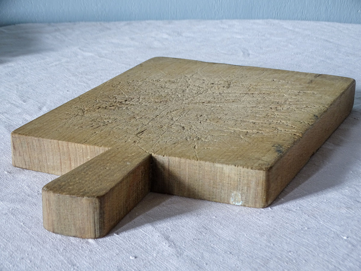 Vintage French wooden chopping board photographed at an angle, showing its long integral handle and worn wooden surface.