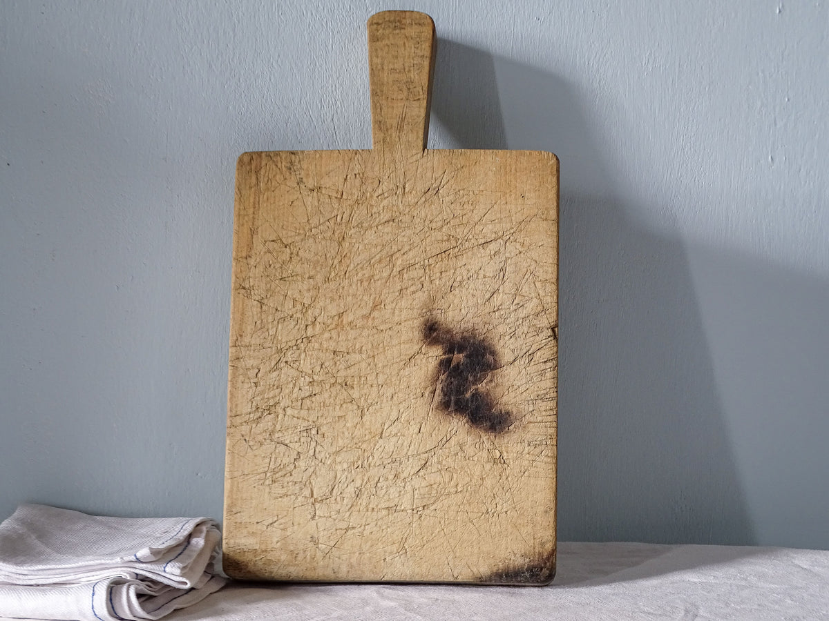 Vintage French wooden cutting board with long handle, pale wood, extensive knife marks and an old scorch mark on one face.