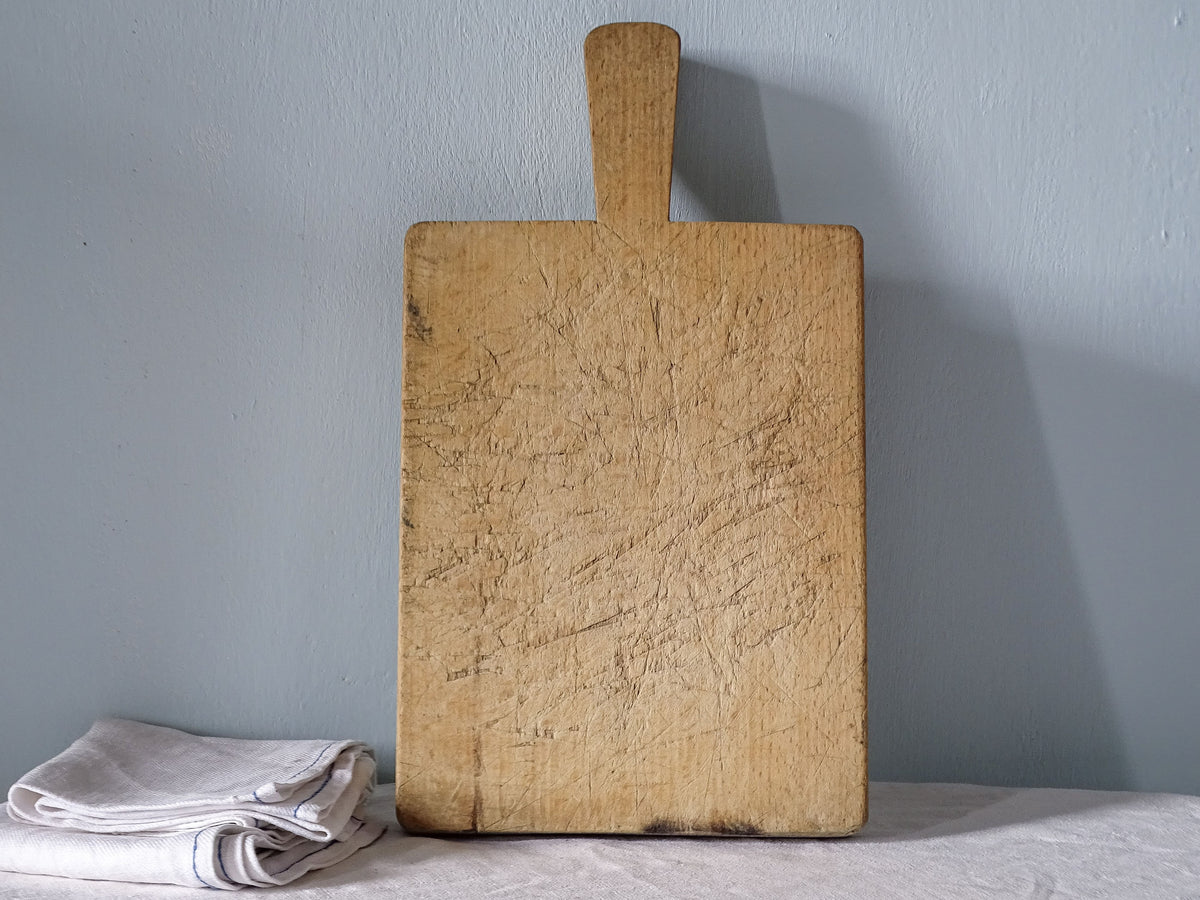 Vintage French wooden cutting board with long handle, rectangular body and heavily knife-marked pale honey-coloured wood.