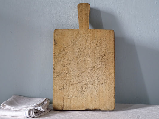 Vintage French wooden cutting board with long handle, rectangular body and heavily knife-marked pale honey-coloured wood.