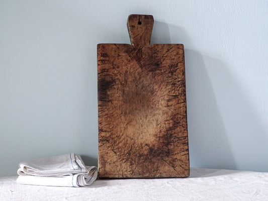 French antique wooden cutting board with a short handle, rich brown patina and heavily marked surface, displayed upright in a rustic kitchen setting.
