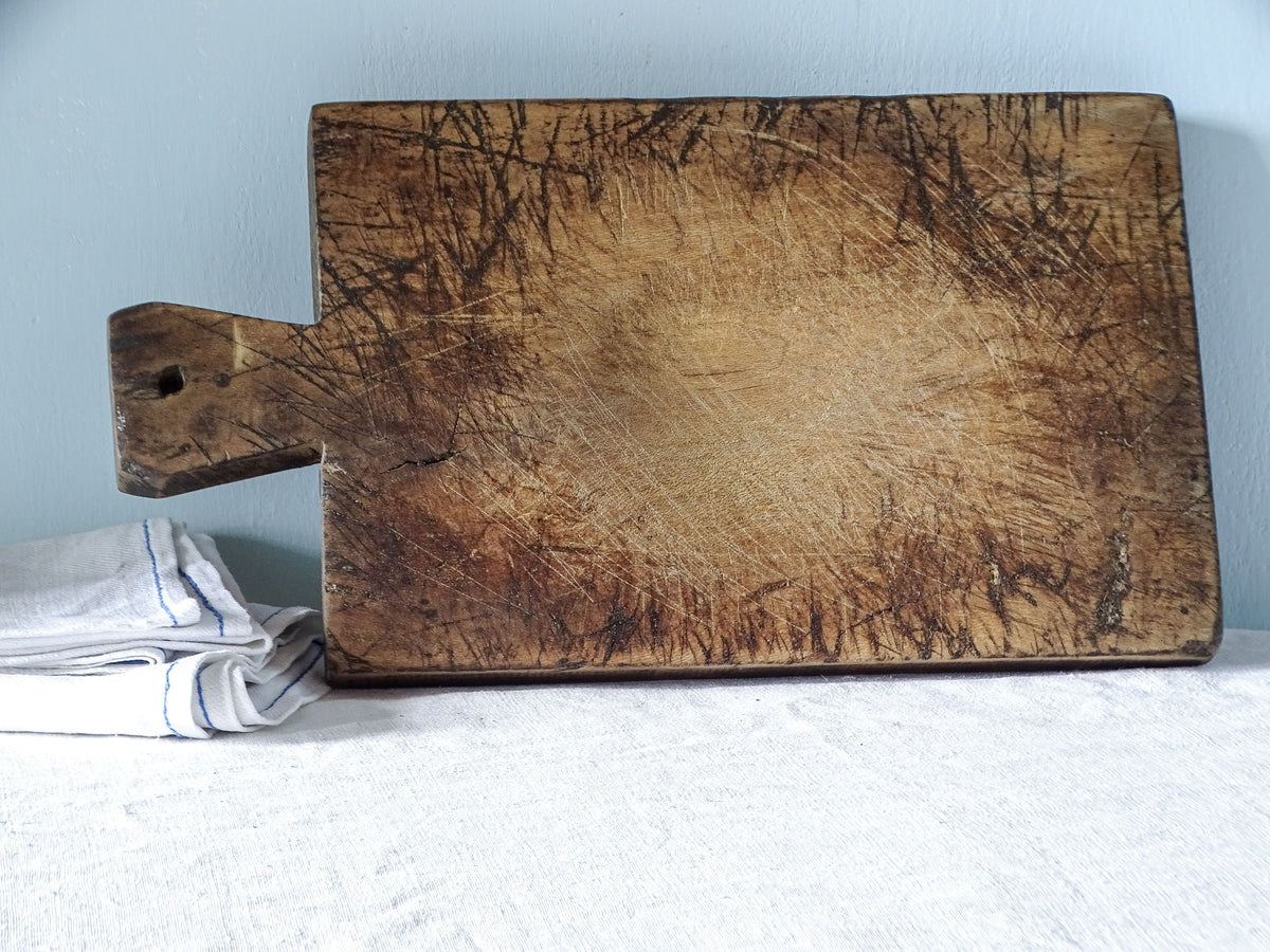 French antique wooden cutting board displayed horizontally, showing its rectangular shape, short handle and heavily scratched surface with a worn centre.
