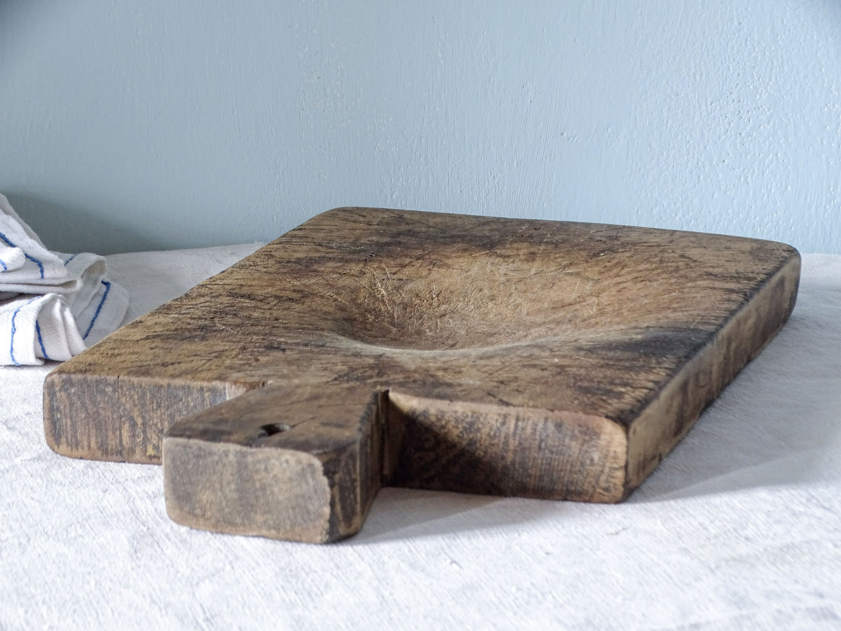 Angled side view of a thick antique wooden chopping board, showing the shallow hollow worn into the centre and the aged handle.