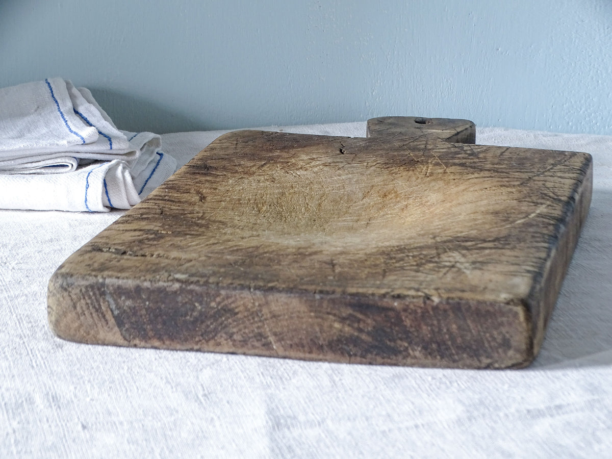 Low-angle view of a French antique wooden cutting board showing its substantial thickness, worn edges and recessed chopping surface.