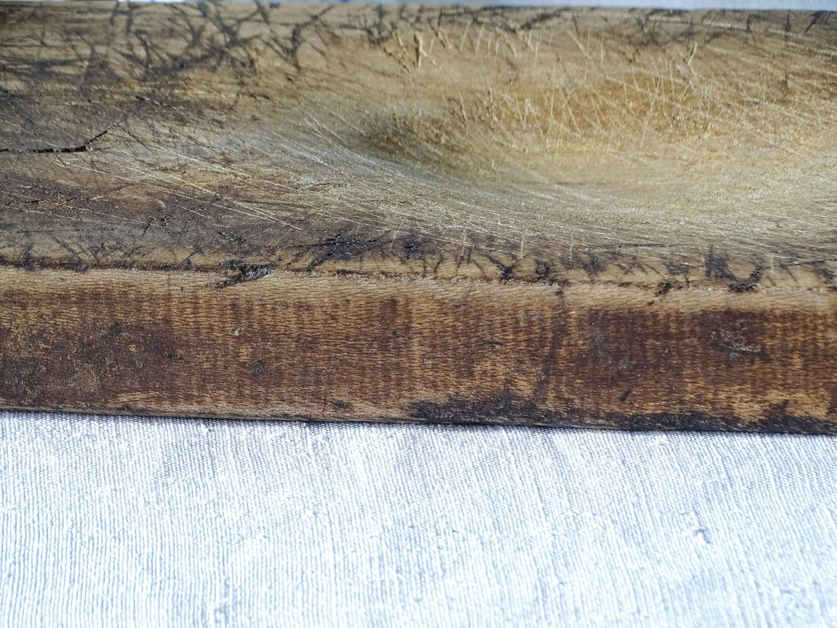 Close-up of the edge of an antique wooden cutting board, showing the aged wood grain, extensive knife marks and dark brown patina.