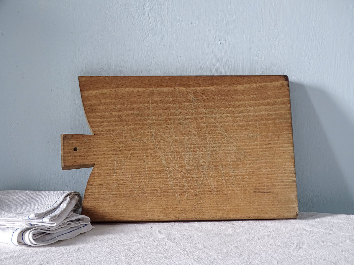 Vintage French rectangular wooden cutting board with squared handle, angled shoulders and warm golden-brown wood covered in fine knife marks.