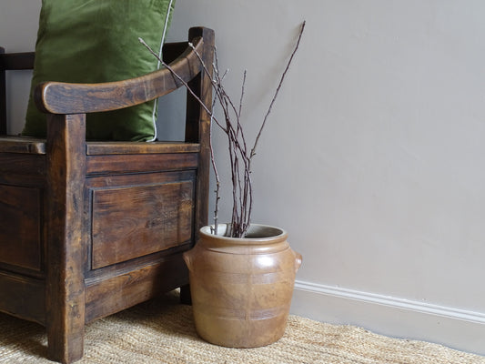 French antique stoneware confit pot with a brown salt glaze and filled with branches. The ceramic jar sits on a natural jute rug and next to an antique wooden seat.