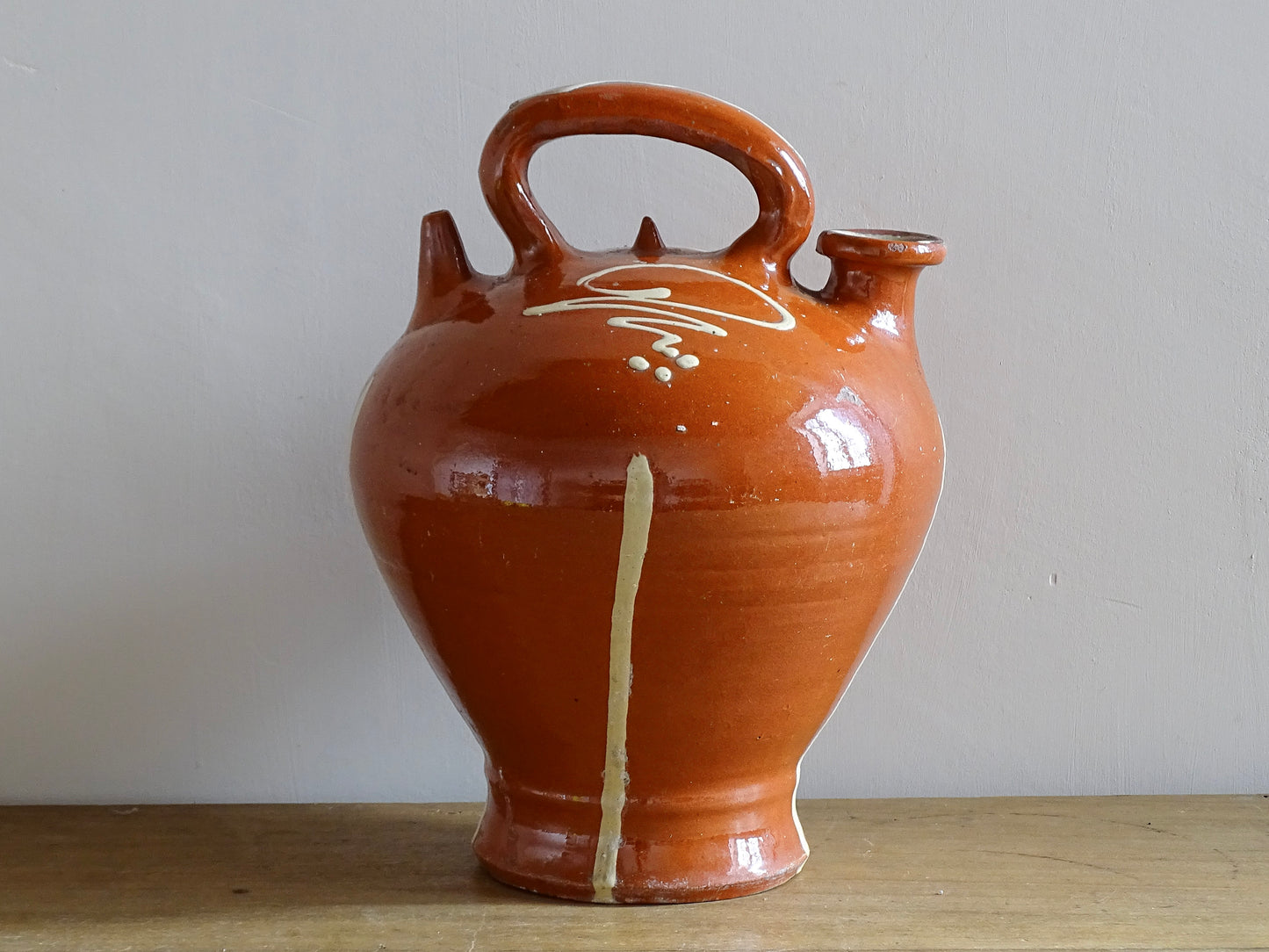 Rustic French antique pottery cruche or gargoulette with a deep red glaze and yellow stripe detailing. The body of this terracotta pitcher is a rounded shape with a handle to the top and two spouts. Glazed pottery water jug for a Provençal style farmhouse table.