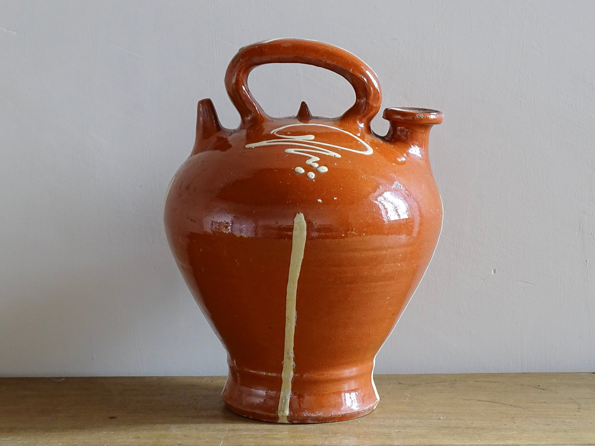 Rustic French antique pottery cruche or gargoulette with a deep red glaze and yellow stripe detailing. The body of this terracotta pitcher is a rounded shape with a handle to the top and two spouts. Glazed pottery water jug for a Provençal style farmhouse table.