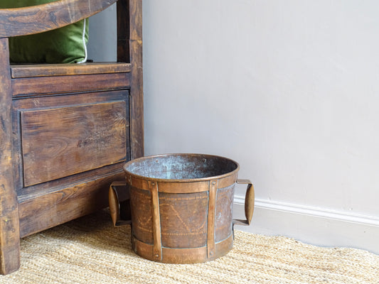 French antique copper water carrier or pot, known as a ferrat. The copper vessel has two handles and sits upon a natural jute rug, alongside an antique wooden seat.