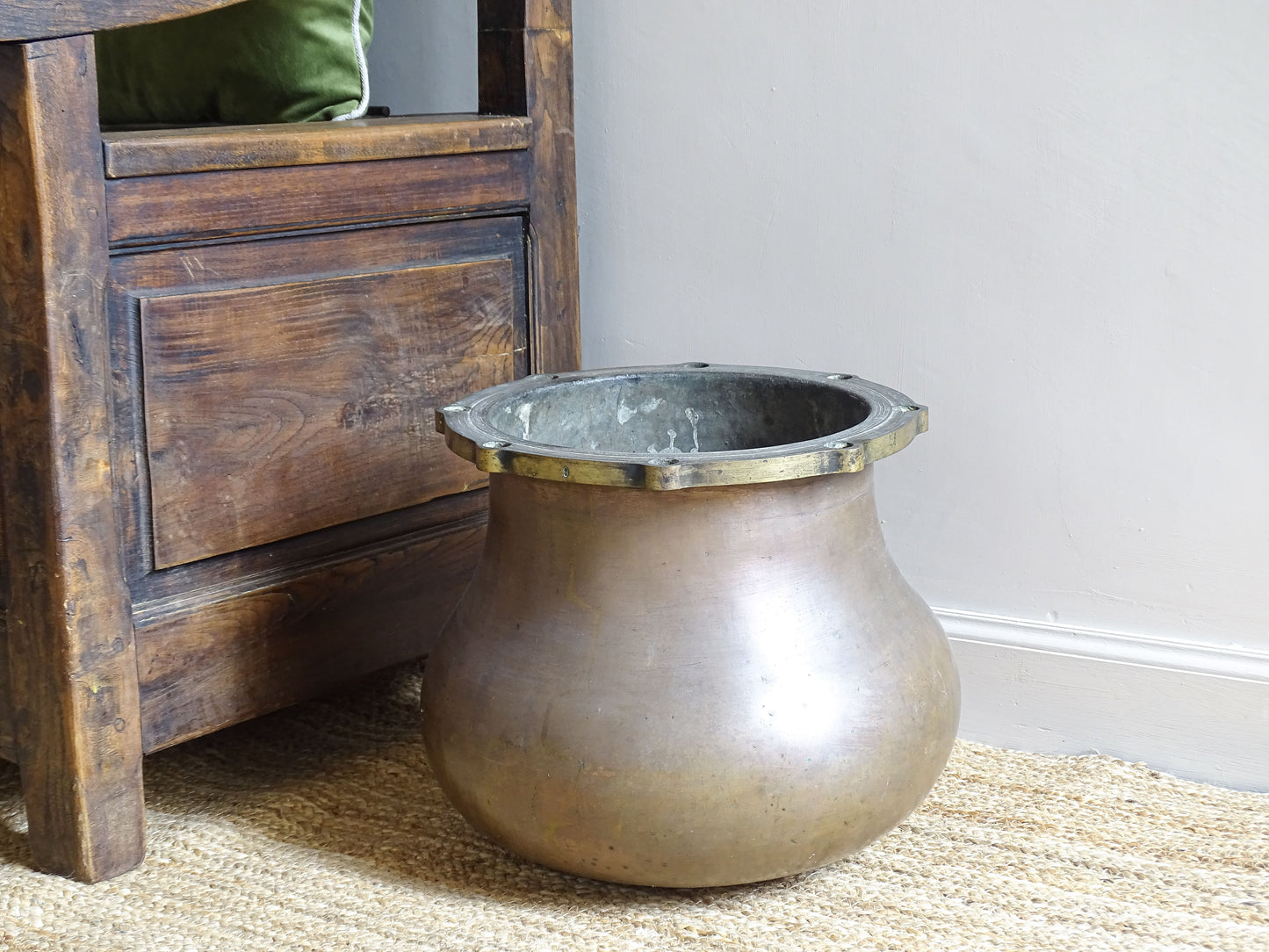 French antique copper pot with brass top, sitting on a natural jute rug and next to an antique wooden chair.