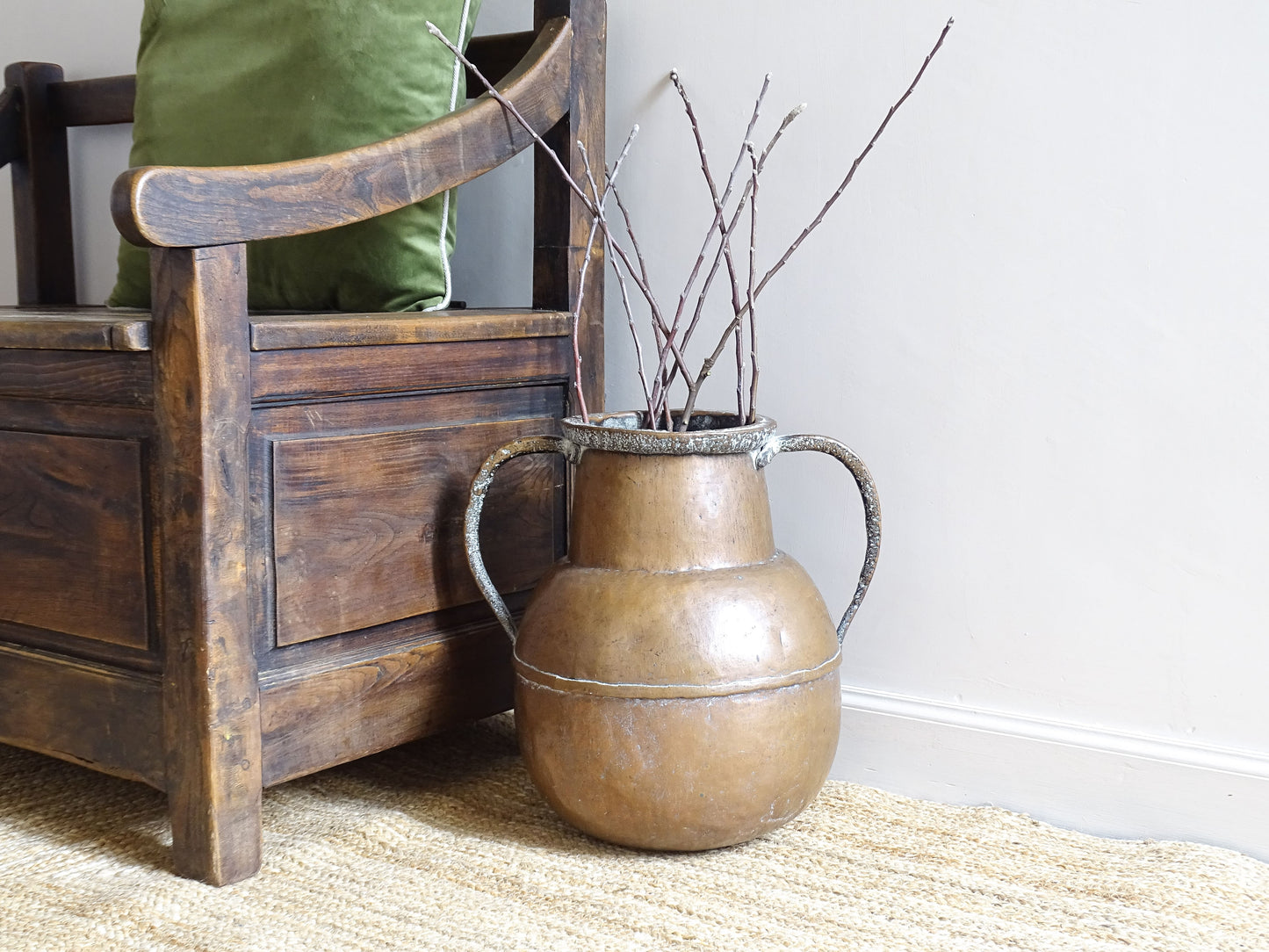French antique copper water pot with two handles and filled with branches. The copper vessel sits on a natural jute rug, alongside an antique wooden seat.