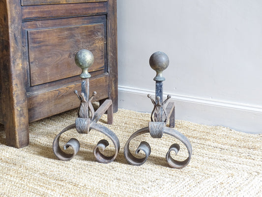 French antique wrought iron andirons with curved base, leaf motifs and brass finials. The fire dogs sit upon a natural jute rug, alongside an antique wooden seat.