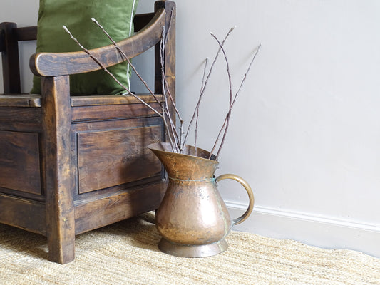 Large French antique copper pitcher with hammered surface, pedestal base and bulbous body. The copper jug sits upon a natural jute rug, alongside an antique wooden seat.