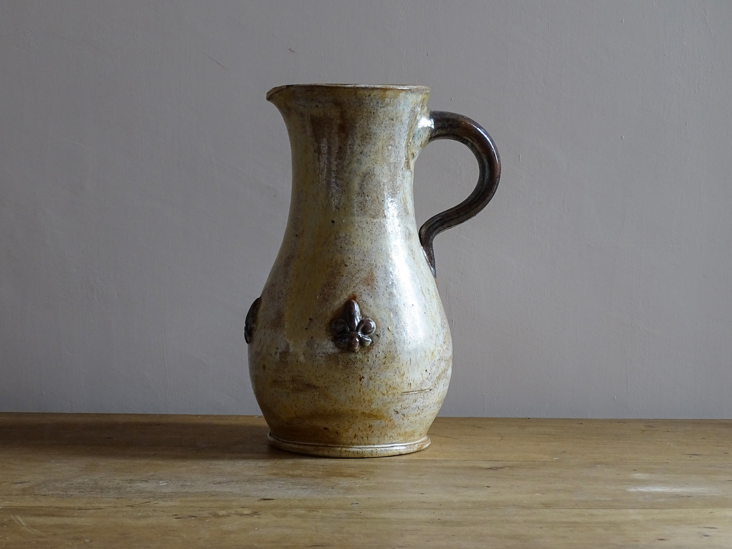 vintage pottery jug with fleur de lys motifs in the style of Roger Guerin. Tall pitcher is glazed in cream, with brown highlights. Display on a sideboard filled with flowers.