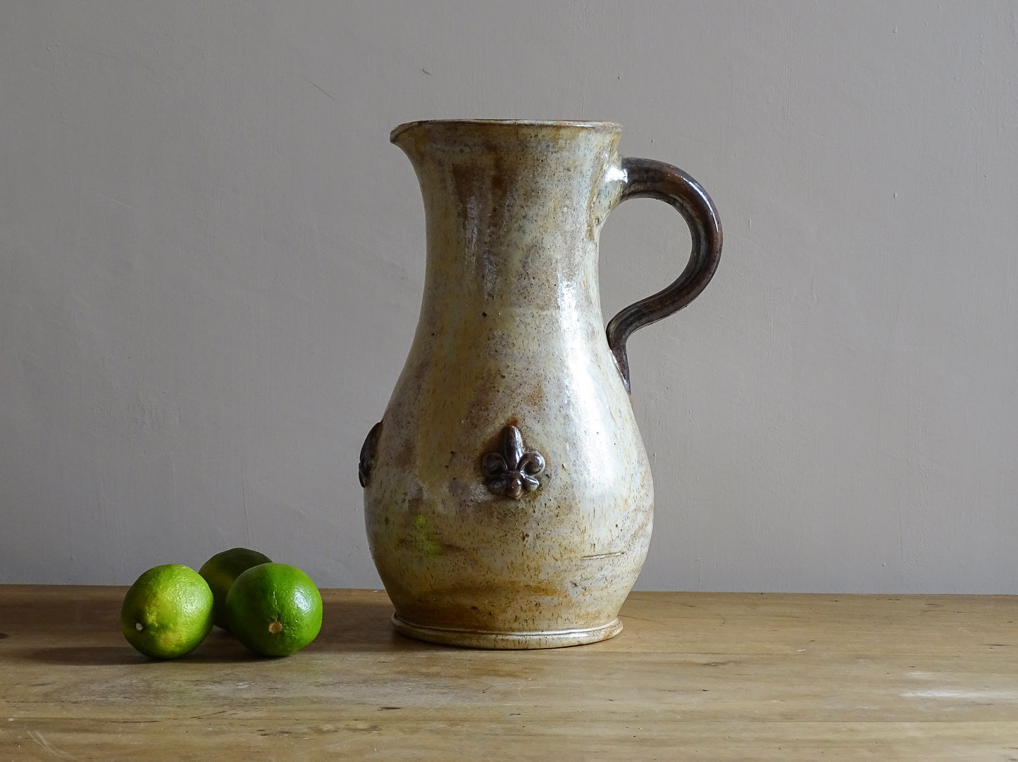 vintage pottery jug with fleur de lys motifs in the style of Roger Guerin. Tall pitcher is glazed in cream, with brown highlights. Display on a sideboard filled with flowers.