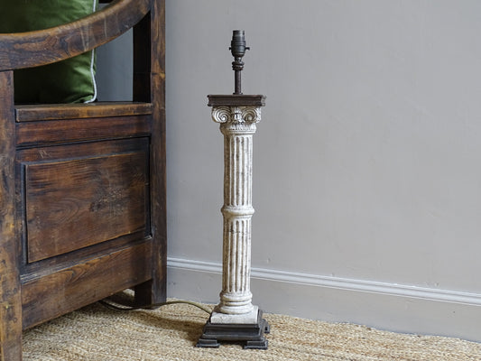 Vintage Corinthian column lamp base sitting on a natural jute rug alongside an antique wooden seat.
