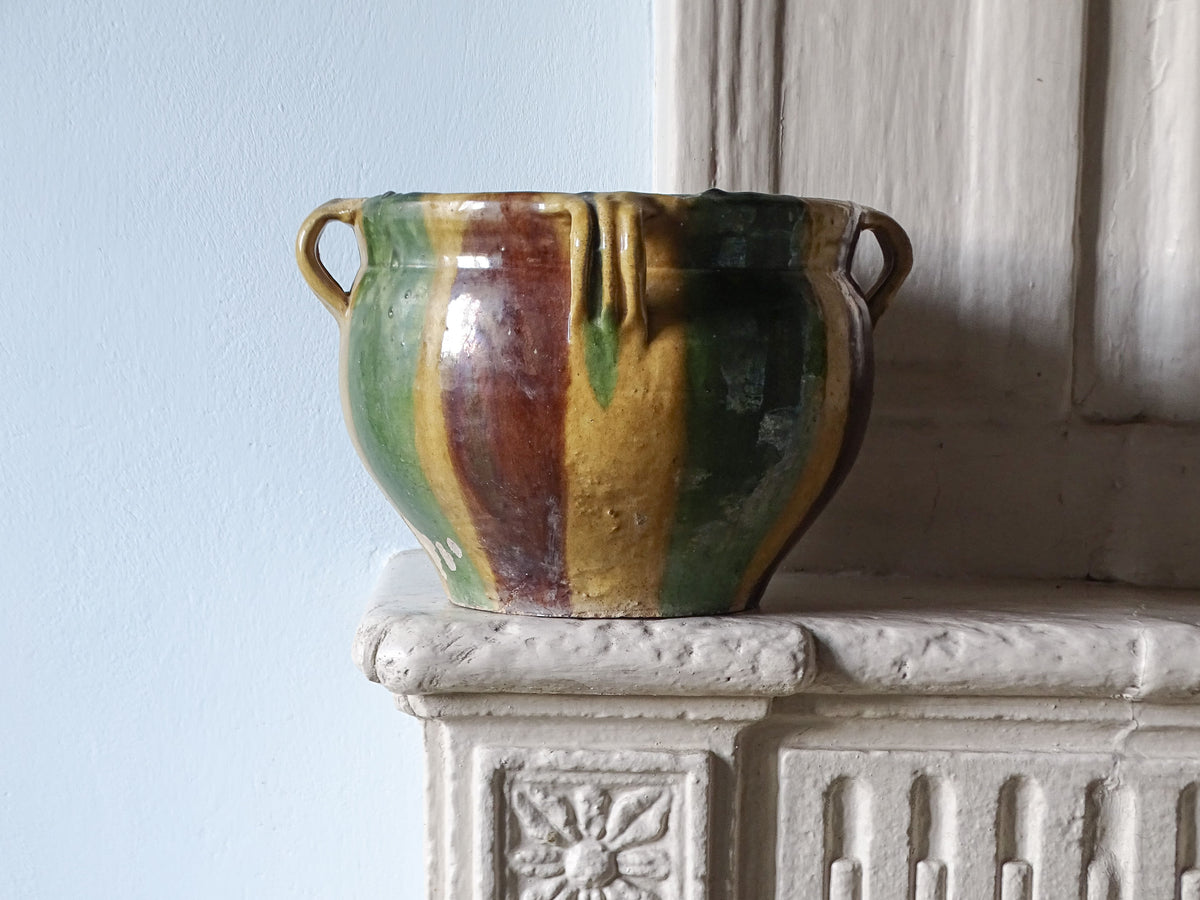 French antique glaze ceramic cache pot or planter with yellow, green and brown glaze, pictured on a stone fireplace next to a pale blue wall. 