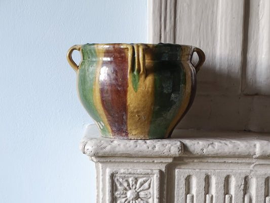 French antique glaze ceramic cache pot or planter with yellow, green and brown glaze, pictured on a stone fireplace next to a pale blue wall. 