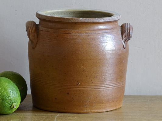 A rustic brown French antique stoneware crock pot with two side handles, sitting on a wooden surface next to two green limes against a neutral off-white wall.