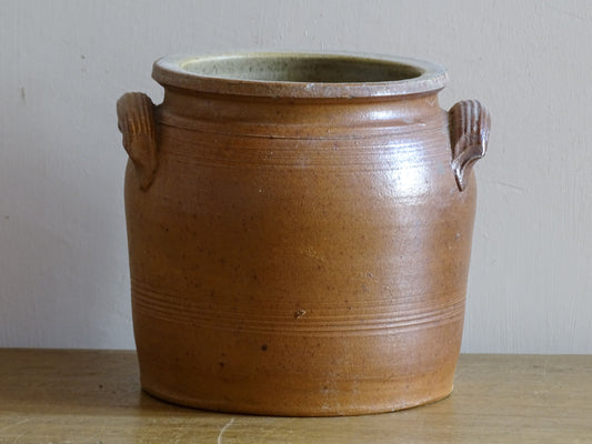 A rustic brown stoneware jar with two side handles, sitting on a wooden surface against an off-white wall. 