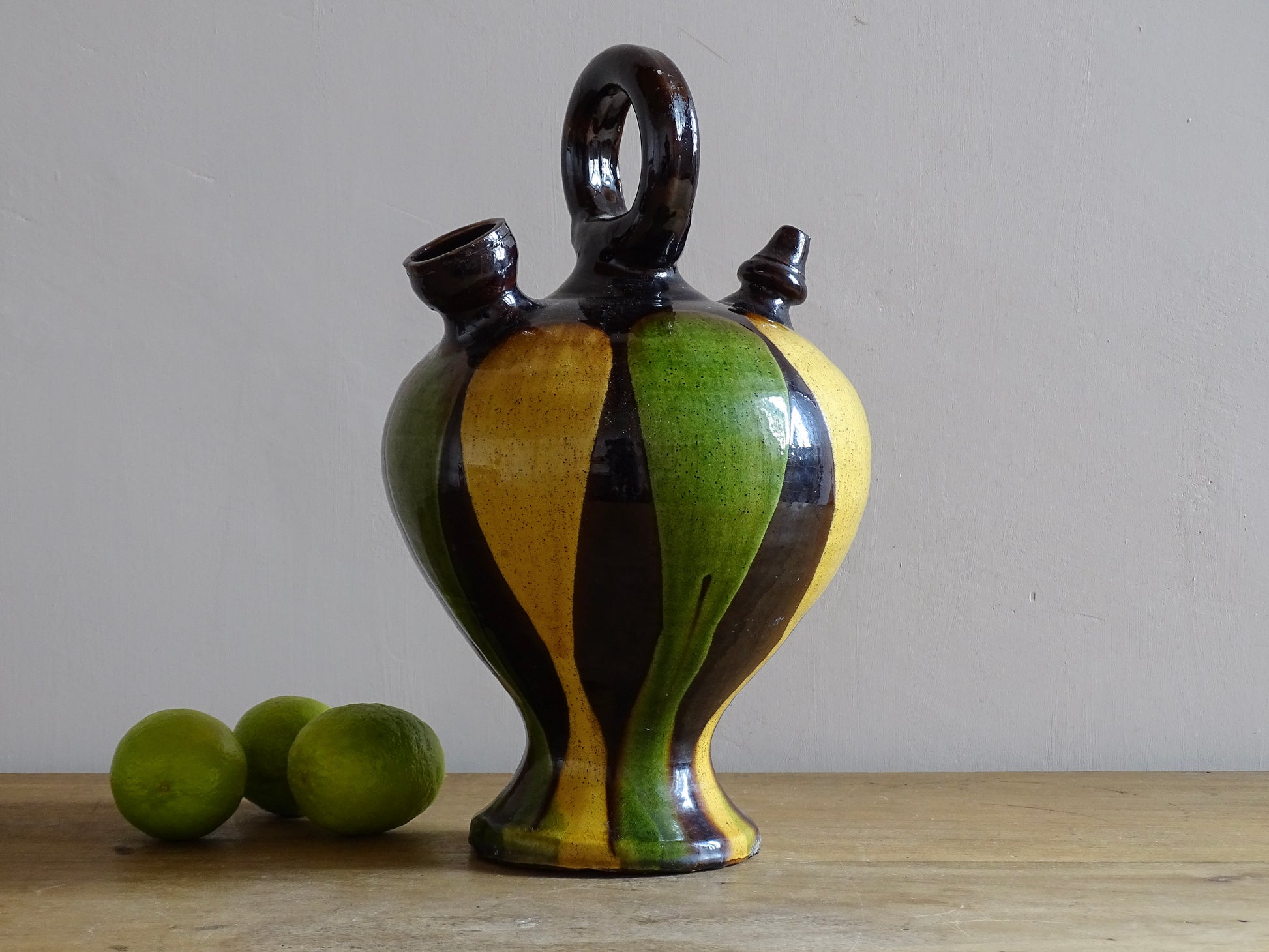 French antique pitcher or gargoulette. Terracotta jug with a green and yellow striped glaze on a dark brown background. It has a curved shape with a round handle to the top and two spouts.
