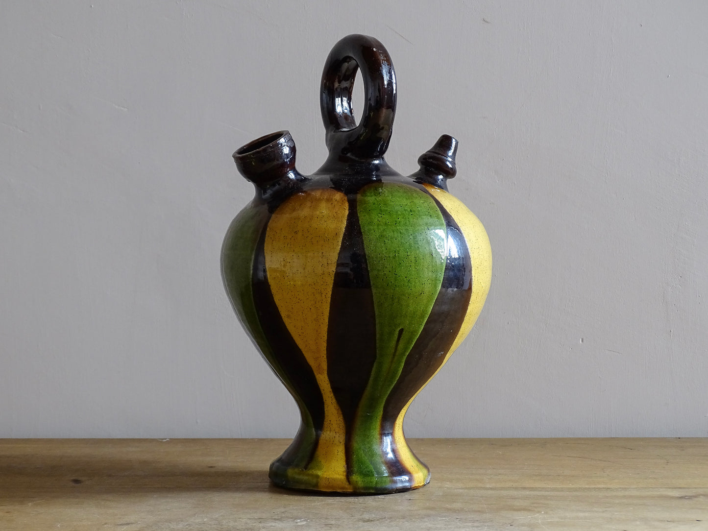 French antique pitcher or gargoulette. Terracotta jug with a green and yellow striped glaze on a dark brown background. It has a curved shape with a round handle to the top and two spouts.