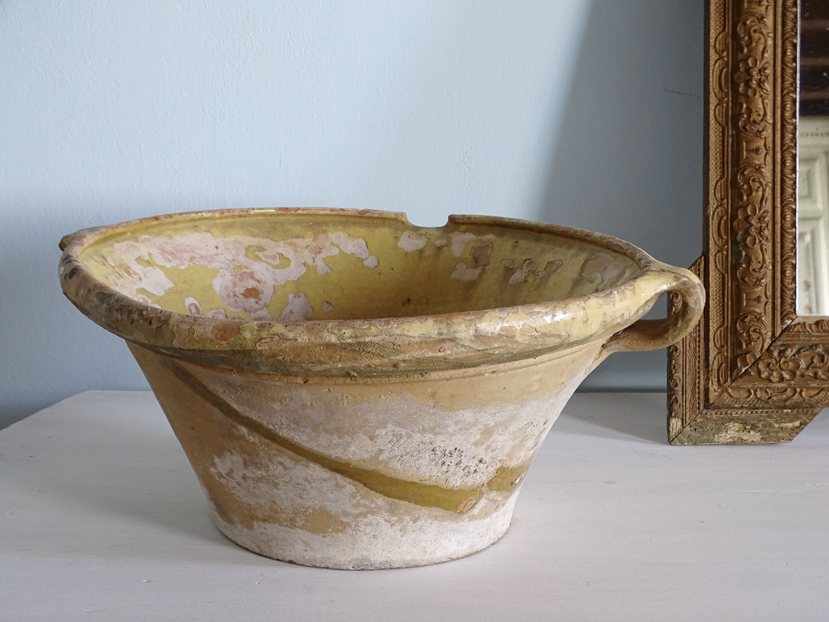 French antique ceramic tian bowl with a worn yellow glaze and loop handle. Pictured on a white table next to a gold mirror in front of a pale blue wall. 