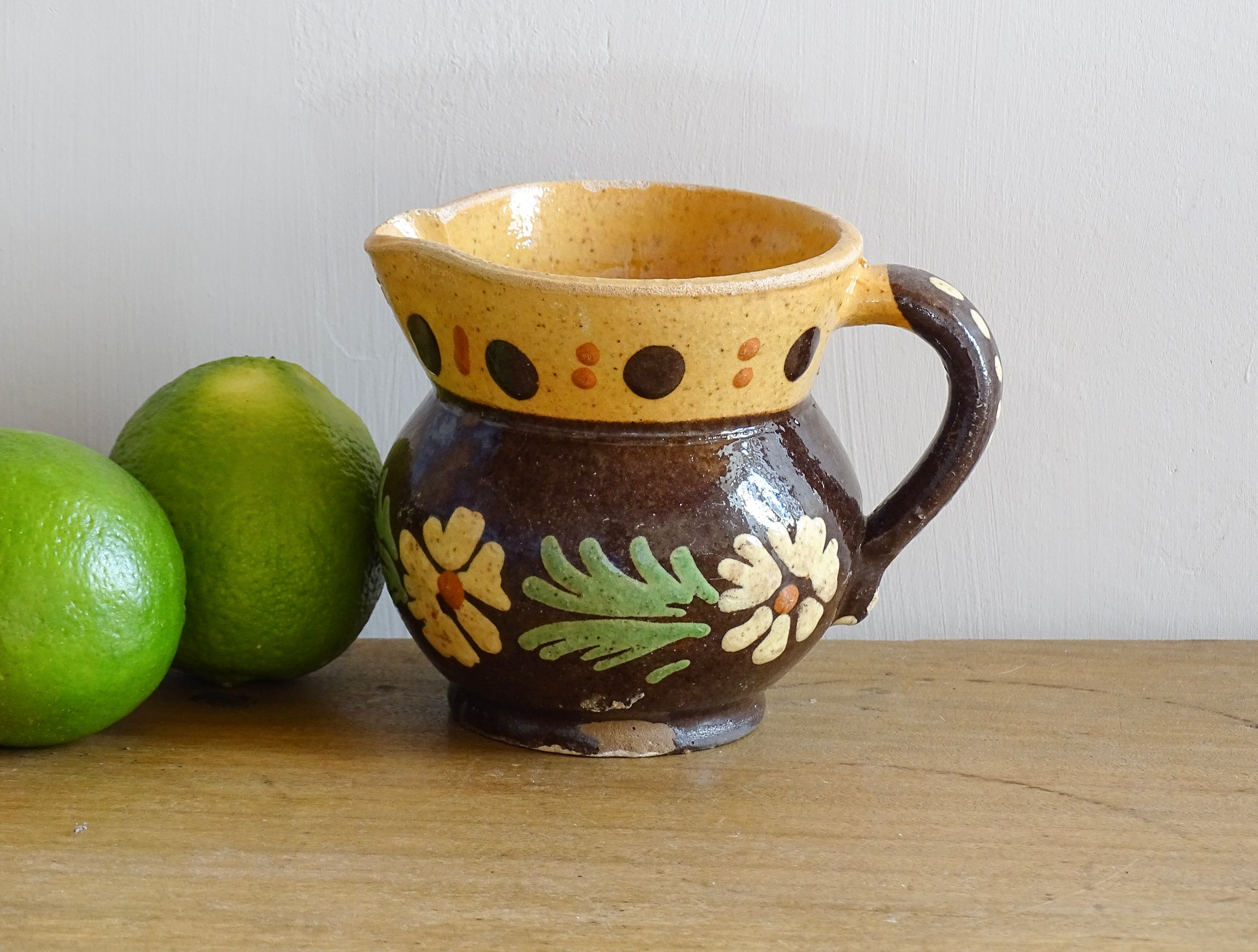 Small French vintage pottery pitcher from Alsace. Ceramic jug with a brown glaze and yellow glazed stripe around the top. It is hand painted with white flowers, green leaves and dots.