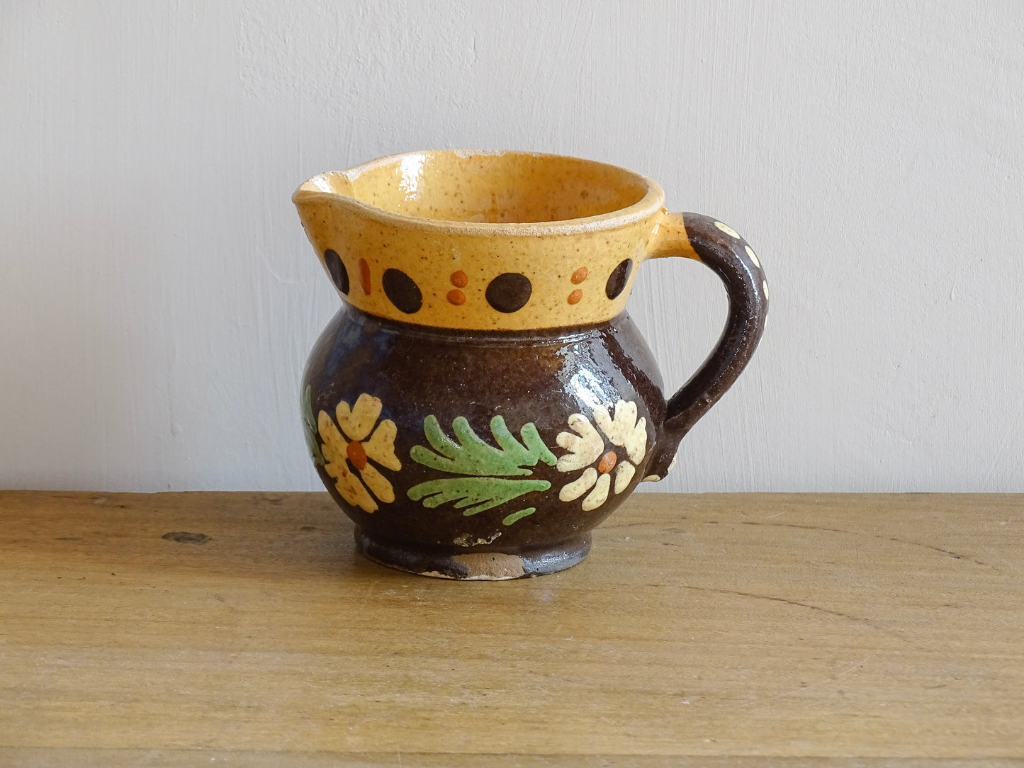 Small French vintage pottery pitcher from Alsace. Ceramic jug with a brown glaze and yellow glazed stripe around the top. It is hand painted with white flowers, green leaves and dots.