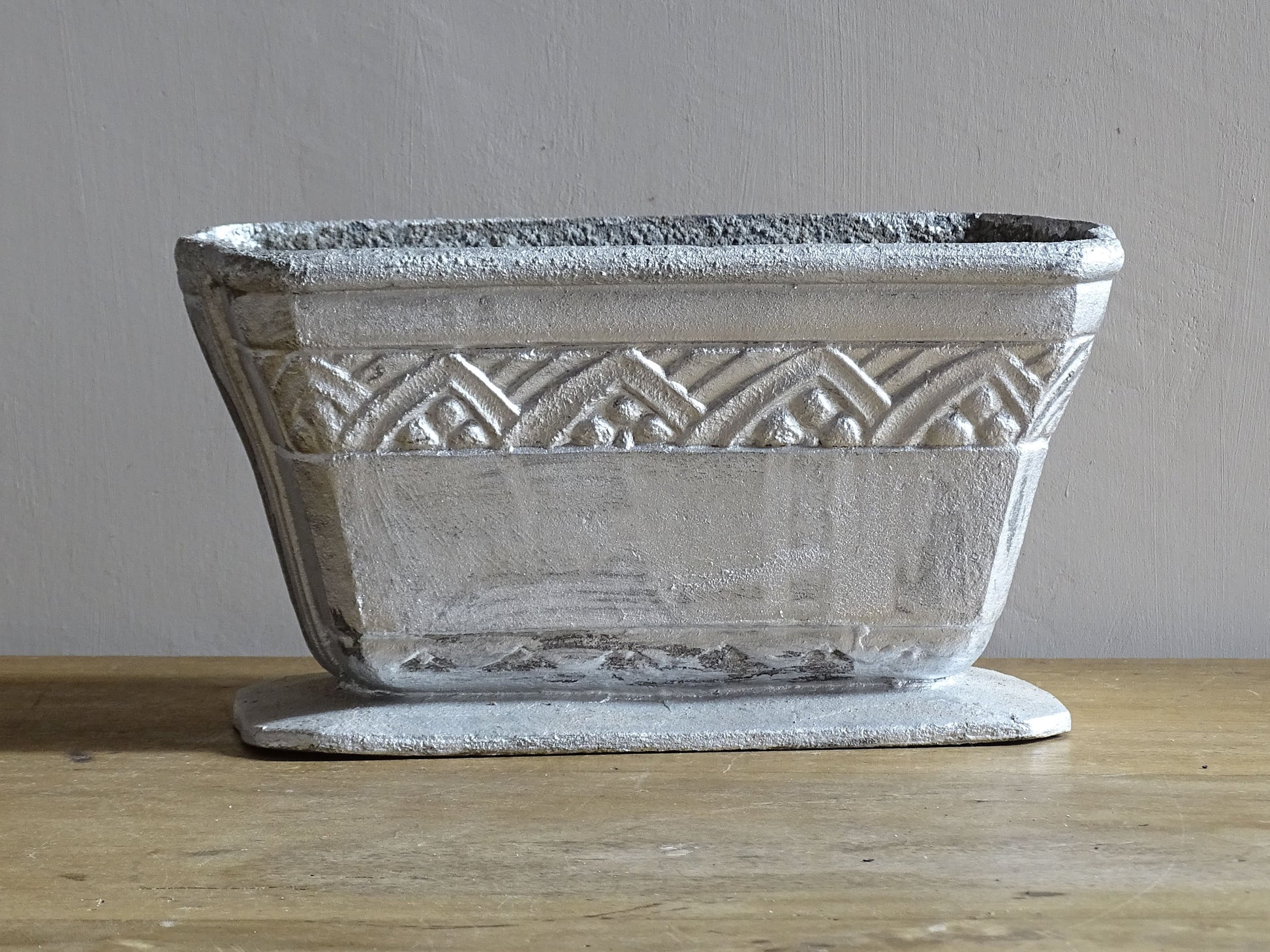 French vintage cast iron planter in silver with a rectangular, flared shape that widens towards the top. It sits on an pedestal-style base. It has a raised geometric border around the upper edge.