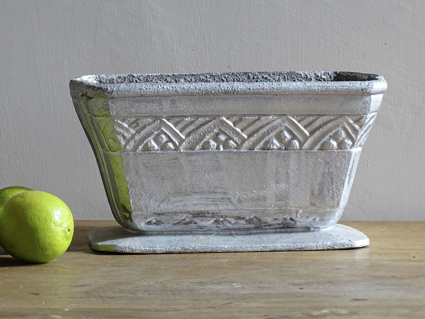 French vintage cast iron planter in silver with a rectangular, flared shape that widens towards the top. It sits on an pedestal-style base. It has a raised geometric border around the upper edge.