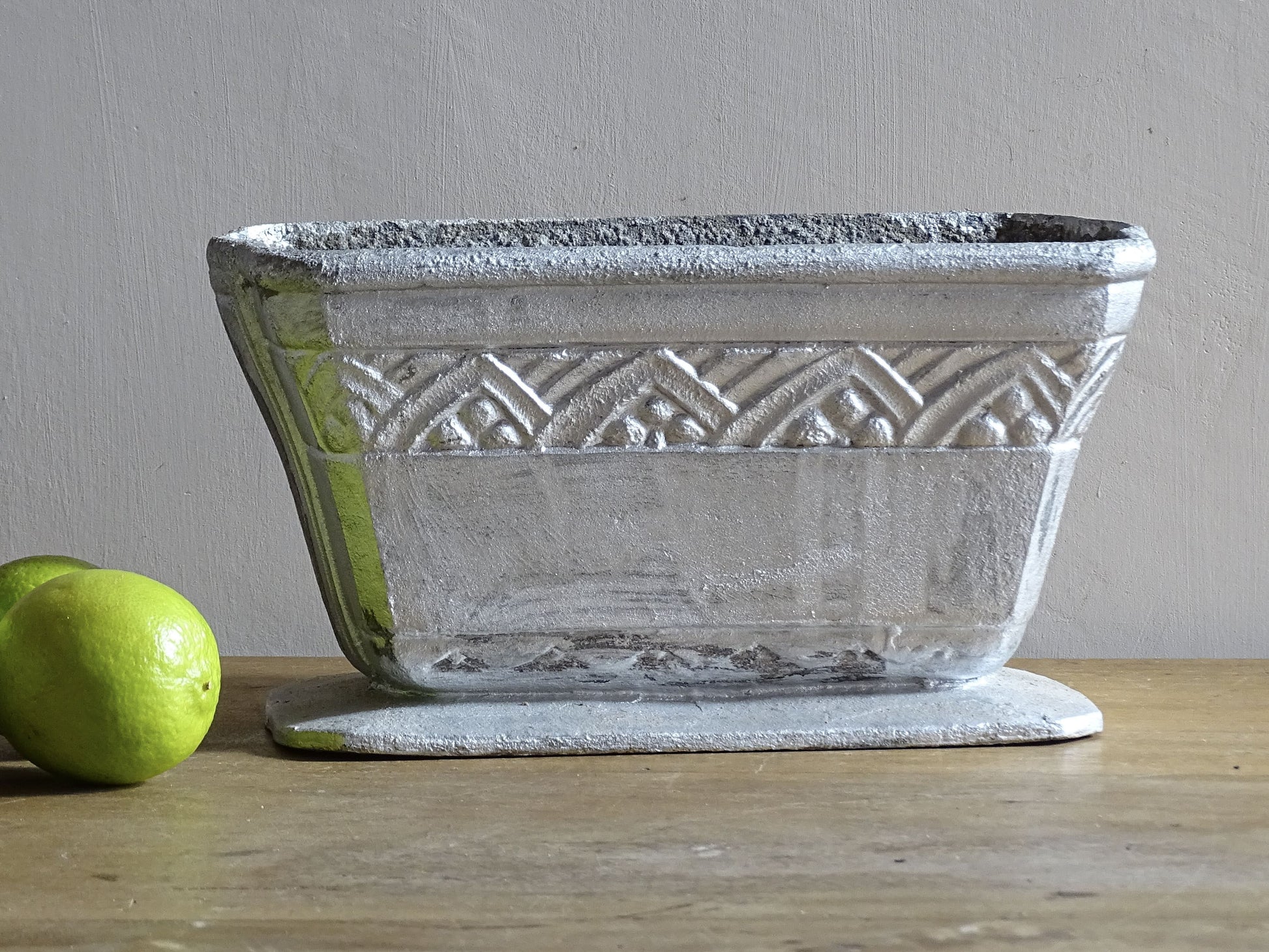 French vintage cast iron planter in silver with a rectangular, flared shape that widens towards the top. It sits on an pedestal-style base. It has a raised geometric border around the upper edge.