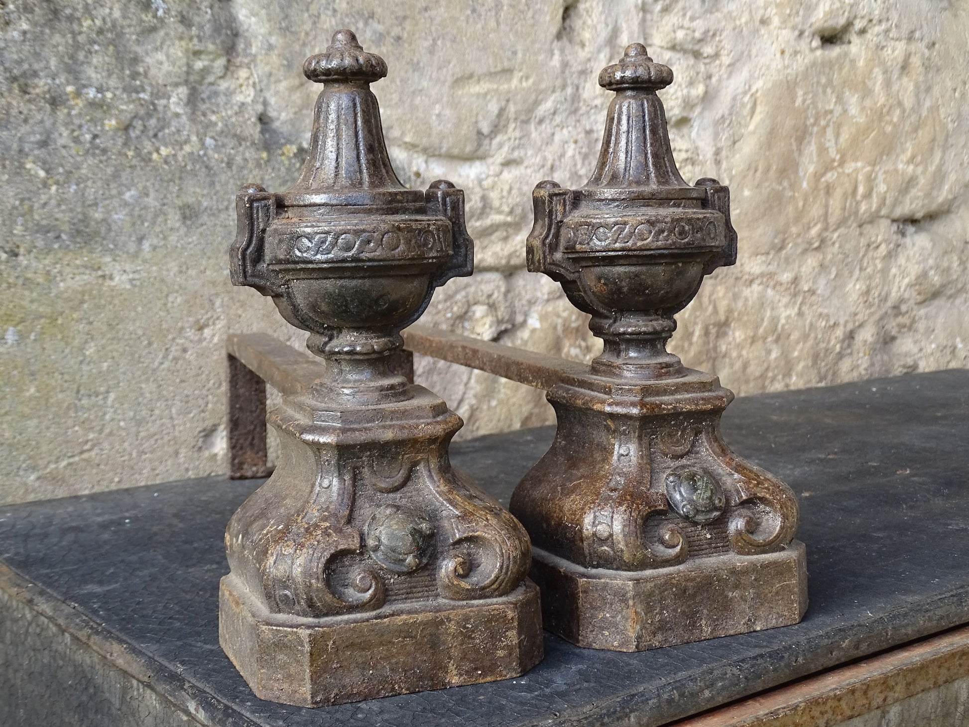Pair of French antique cast iron andirons or fire dogs with a classical style. Each andiron is topped with an urn-shaped finial, accented by moulded details and a fluted form that evokes neoclassical architectural design. The bases are decorated with scrolling motifs and small medallions. 