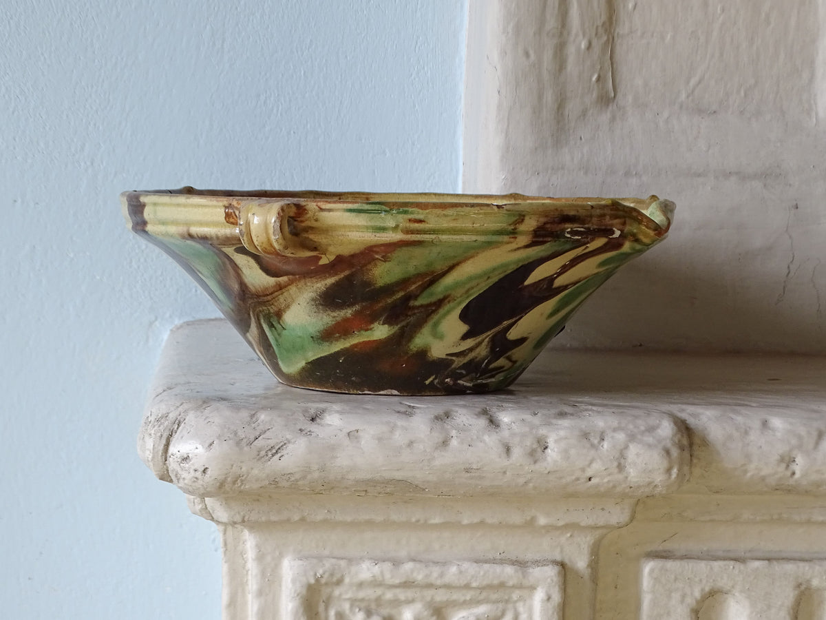 French antique ceramic bowl in green and yellow marble glaze with handles and spout, pictured on a country house fireplace