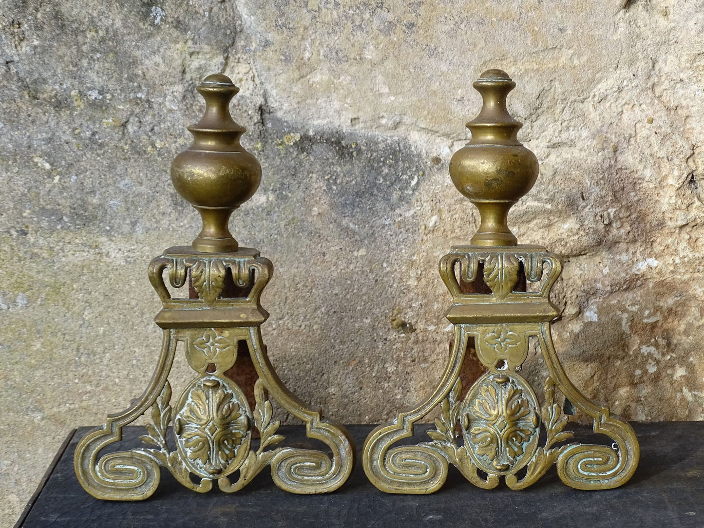 Pair of French antique brass andirons or fire dogs in a Louis XVI revival style. Each andiron is topped with a turned baluster finial. The pierced front plates display an ornate composition of acanthus leaves, scrollwork, and a central oval cartouche framed by laurel branches, typical of neoclassical design.