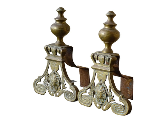 Pair of French antique brass andirons or fire dogs in a Louis XVI revival style. Each andiron is topped with a turned baluster finial. The pierced front plates display an ornate composition of acanthus leaves, scrollwork, and a central oval cartouche framed by laurel branches, typical of neoclassical design.