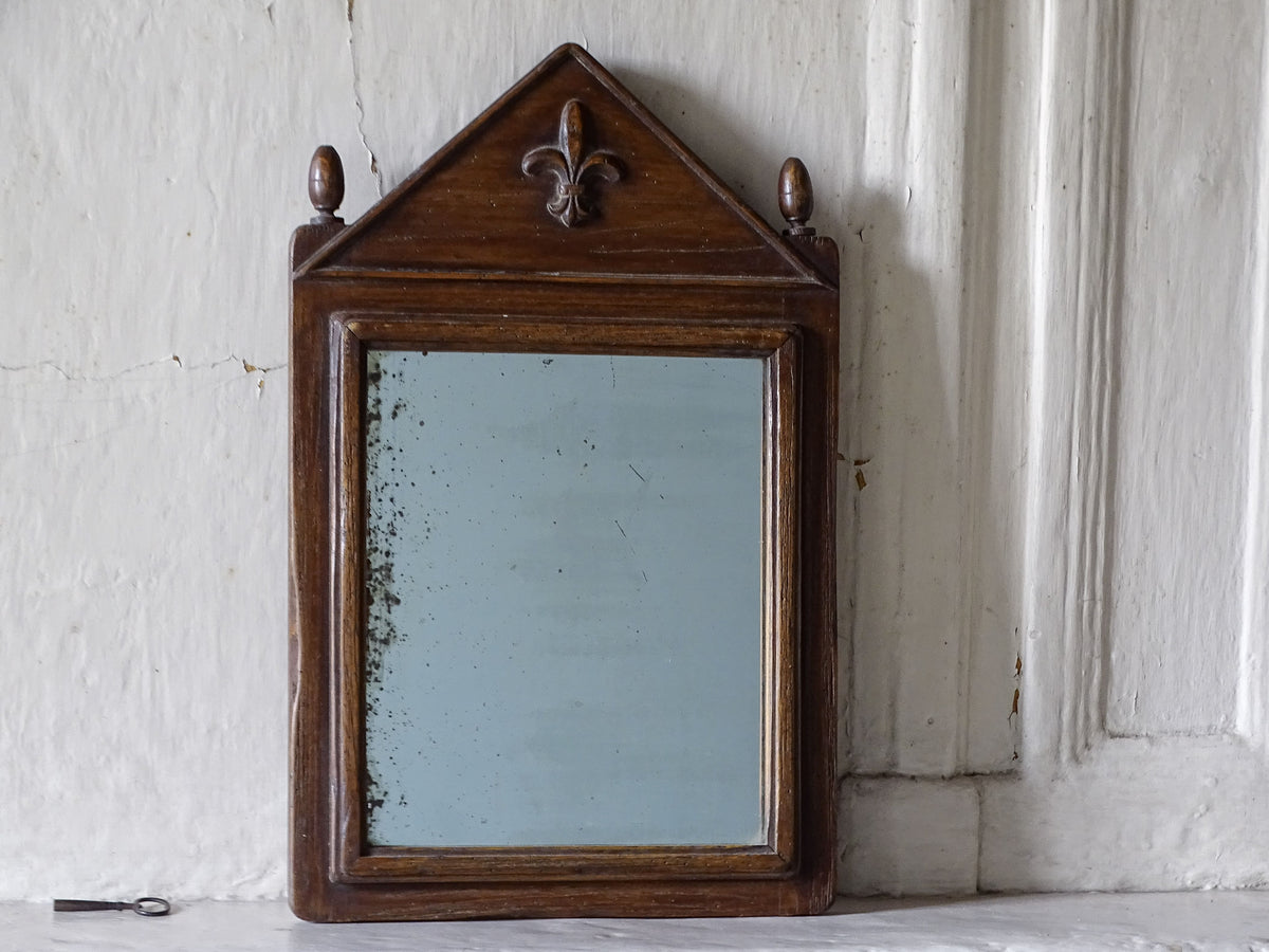 French antique wooden mirror with finials and fleur de lys pediment, displayed on a country house fireplace mantel