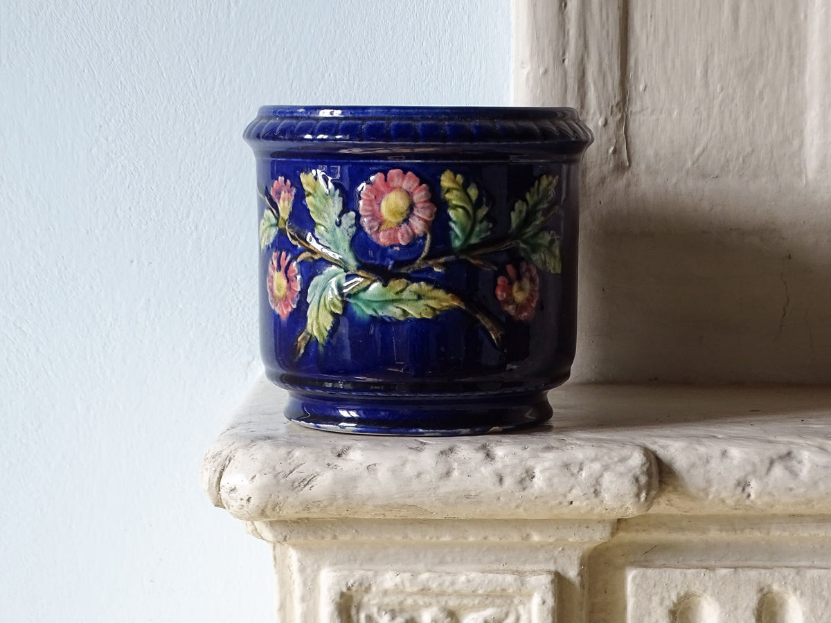 A French antique cobalt blue ceramic planter with hand-painted pink and green floral motifs, displayed on a textured white stone mantelpiece against a light blue wall.