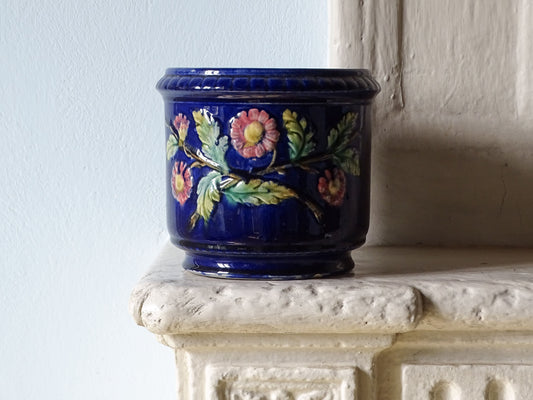 A French antique cobalt blue ceramic planter with hand-painted pink and green floral motifs, displayed on a textured white stone mantelpiece against a light blue wall.