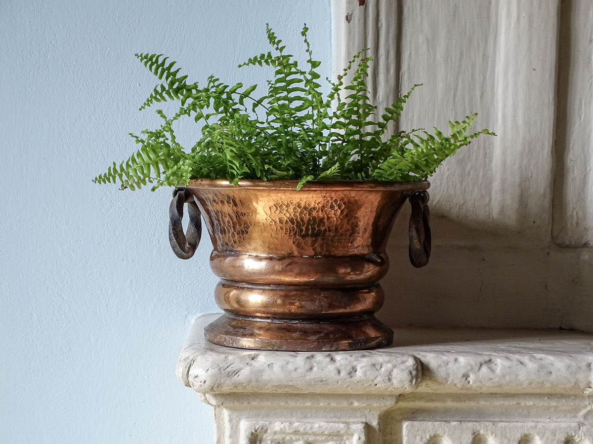 French antique hammered copper planter with wrought iron handles and a fern. Pictured on a country house fireplace next to a pale blue wall