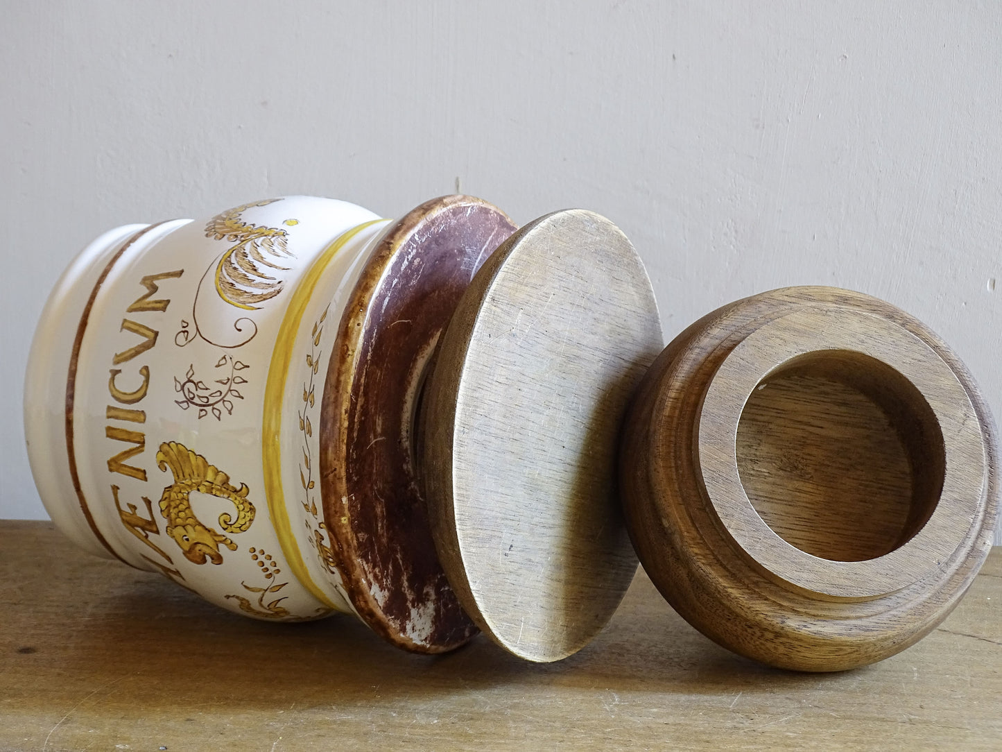 A vintage ceramic apothecary jar with hand-painted decoration and gilt lettering, with a wooden lid and base. Inspired by traditional European pharmacy vessels.