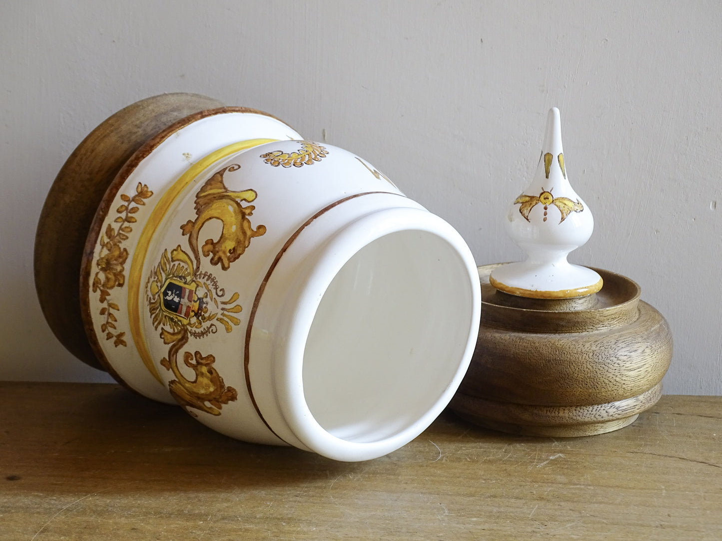 A vintage ceramic apothecary jar with hand-painted decoration and gilt lettering, with a wooden lid and base. Inspired by traditional European pharmacy vessels.