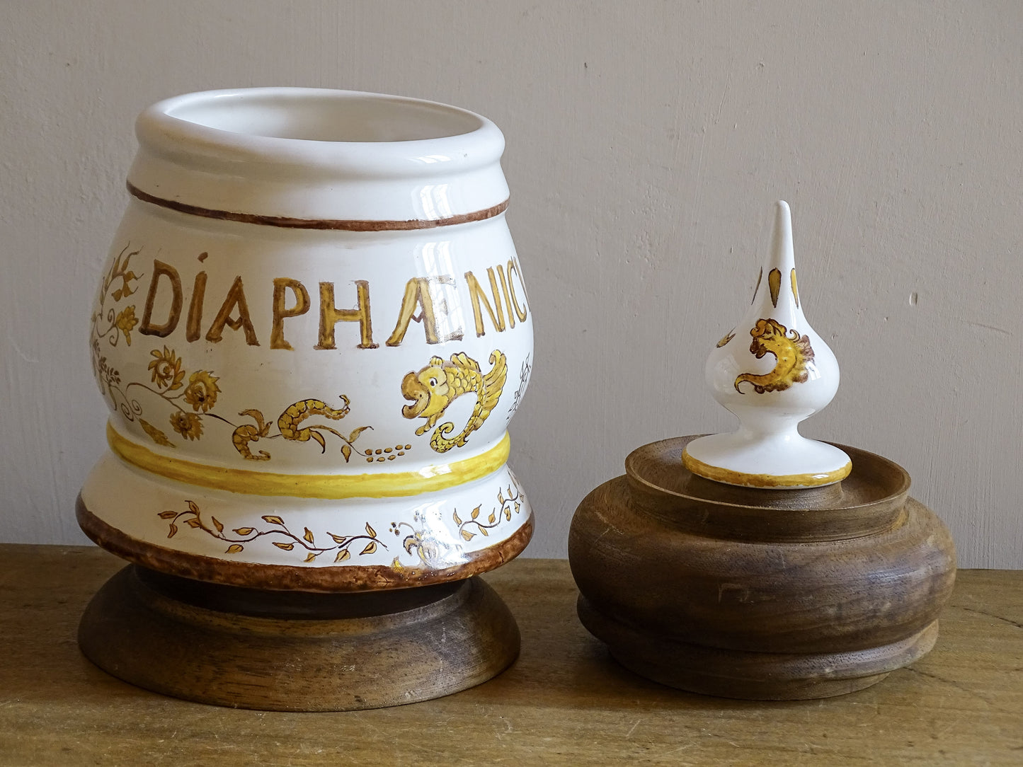 A vintage ceramic apothecary jar with hand-painted decoration and gilt lettering, with a wooden lid and base. Inspired by traditional European pharmacy vessels.