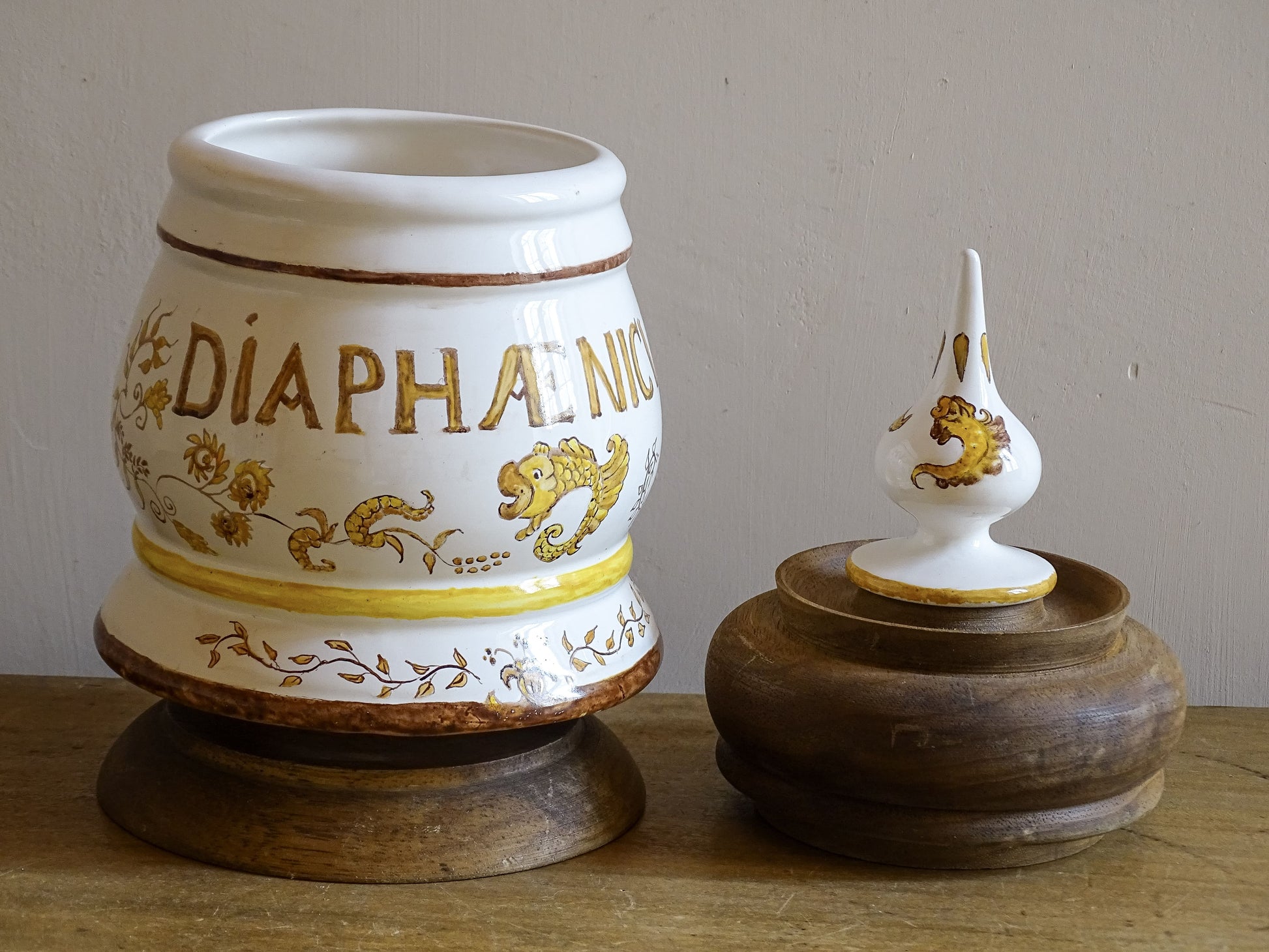A vintage ceramic apothecary jar with hand-painted decoration and gilt lettering, with a wooden lid and base. Inspired by traditional European pharmacy vessels.